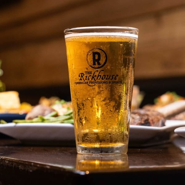 A cold glass of beer with The Rickhouse logo, condensation on the glass, and food in the background.