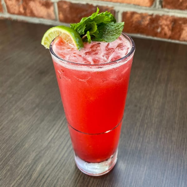 A vibrant red cocktail with ice, garnished with a lime wedge and fresh mint, on a wooden table.