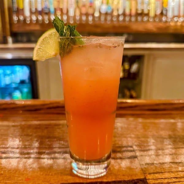 A vibrant orange cocktail with a lime wedge and mint garnish sits on a wooden bar.