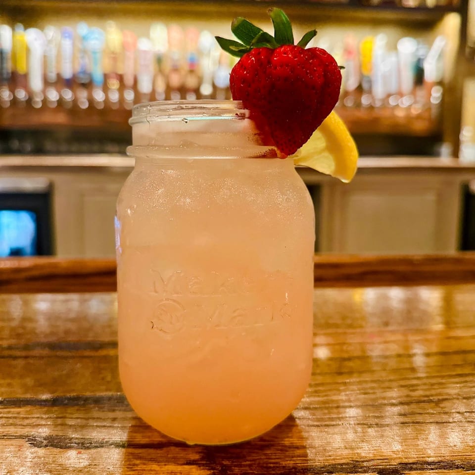 A refreshing strawberry lemonade in a mason jar with a lemon slice on a wooden bar.